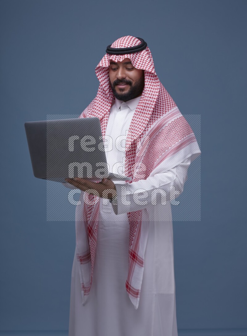 A man standing  with a laptop on Blue Background wearing Saudi Thob and Shomag