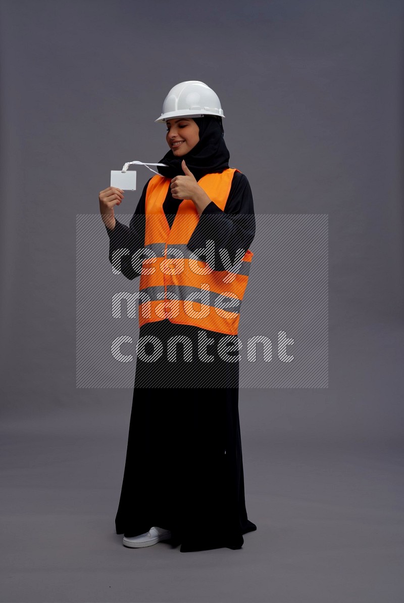 Saudi woman wearing Abaya with engineer vest with neck strap employee badge standing interacting with the camera on gray background