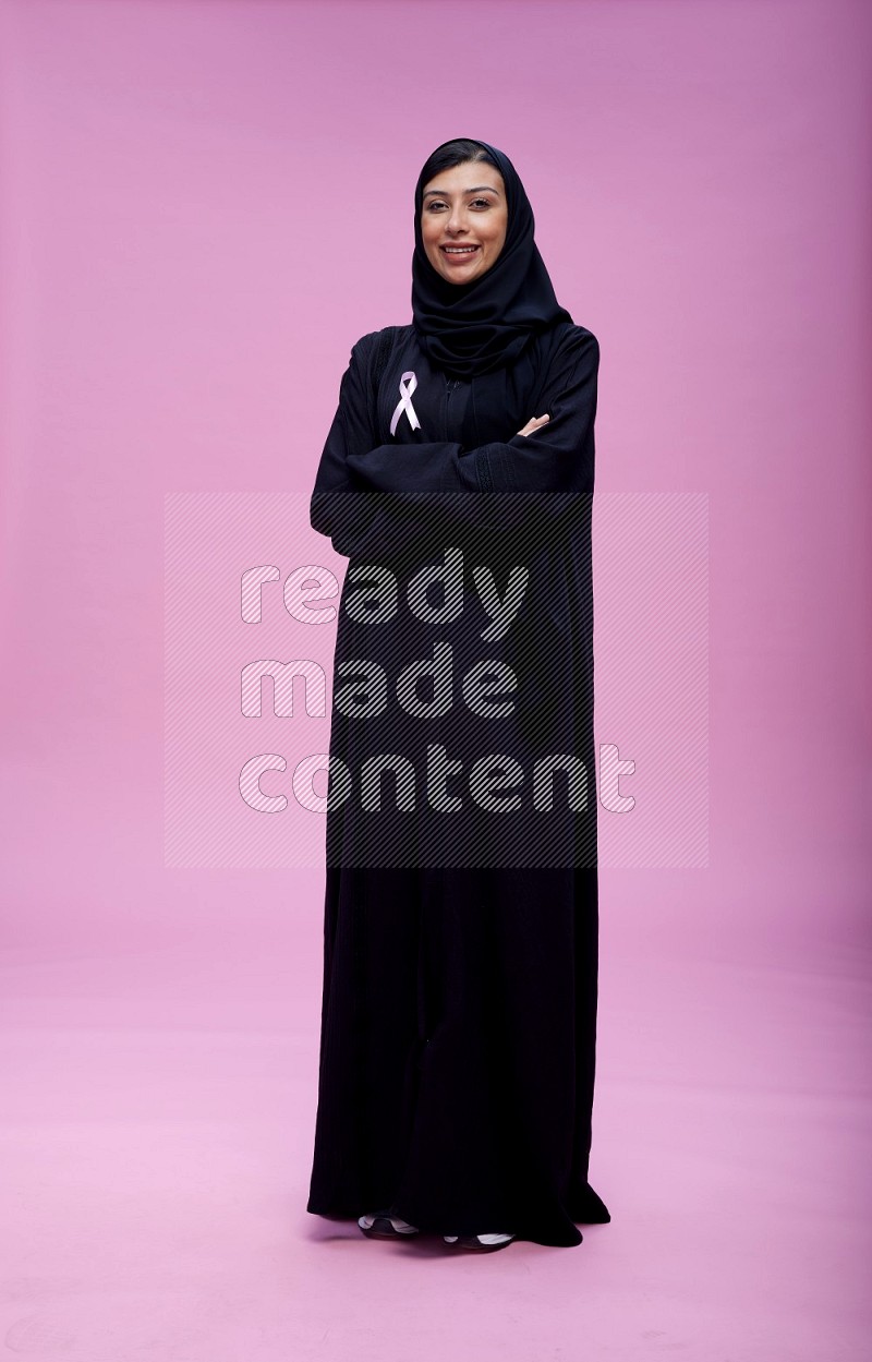 Saudi woman wearing pink ribbon on Abaya standing crossed arms on pink background
