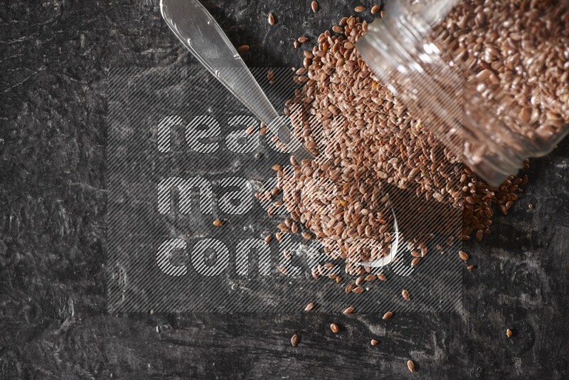 وعاء زجاجي مقلوب ممتلئ بحبوب بذر الكتان والحبوب متناثرة منه مع ملعقة معدنية ممتلئة علي خلفية سوداء