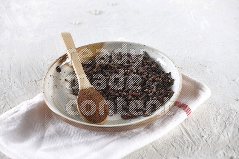 A Pottery plate full of whole cloves and a wooden spoon full of cloves powder in it on a textured white background