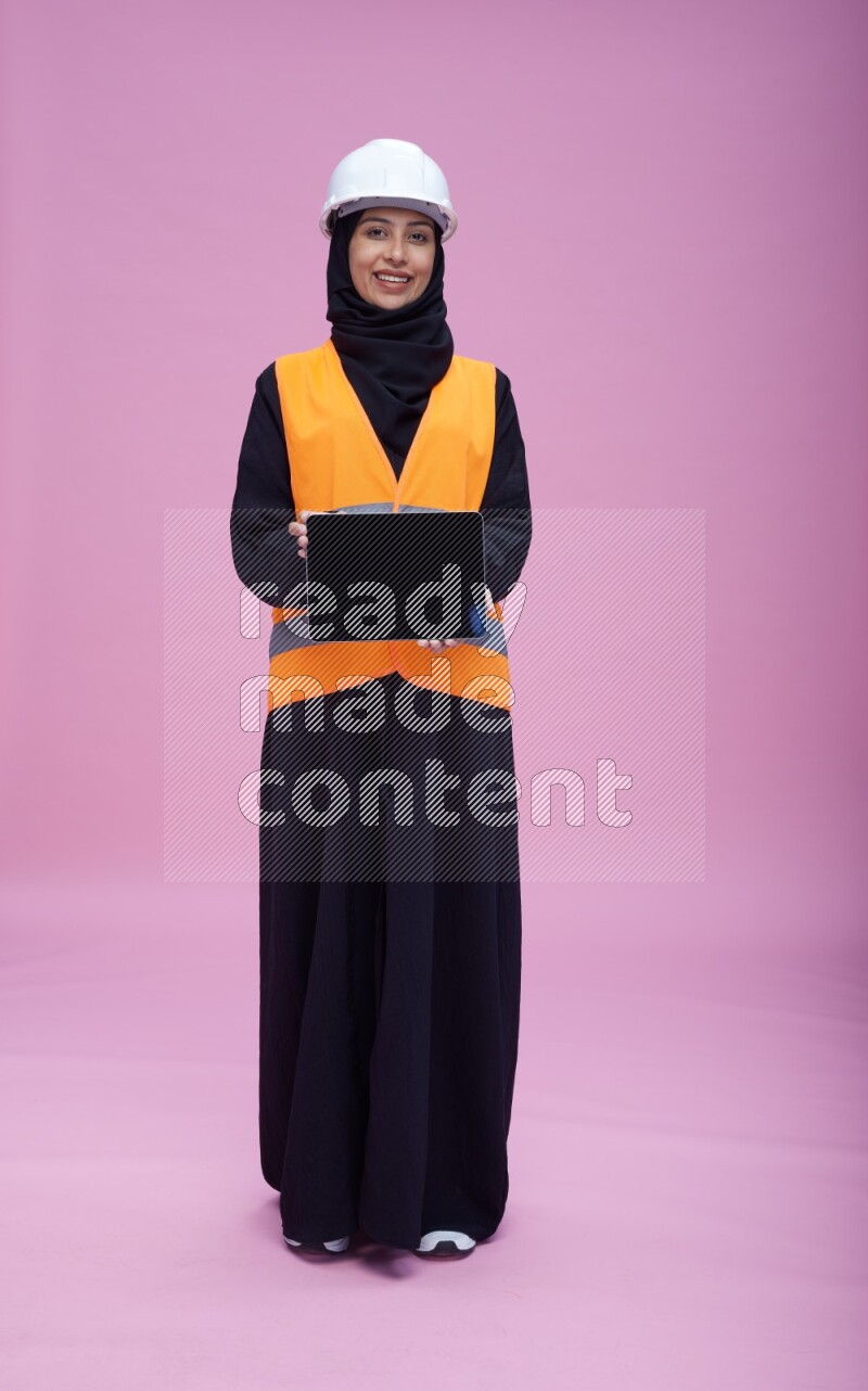 Saudi woman wearing Abaya with engineer vest and helmet standing showing tablet to camera on pink background
