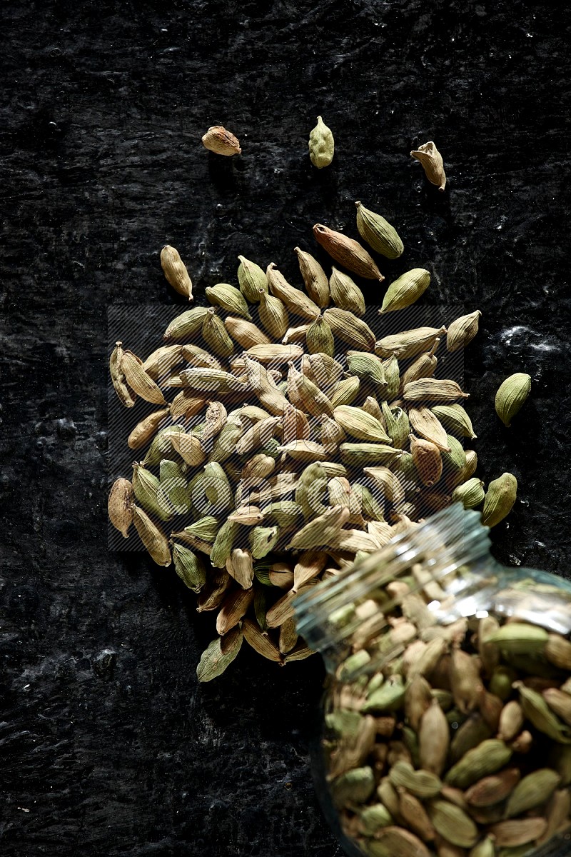A flipped glass spice jar full of cardamom seeds on textured black flooring