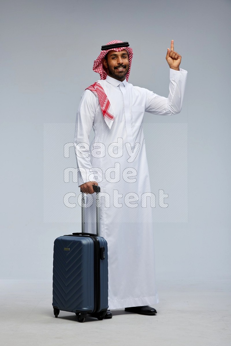 Saudi man wearing Thob and shomag standing holding Travel bag on Gray background