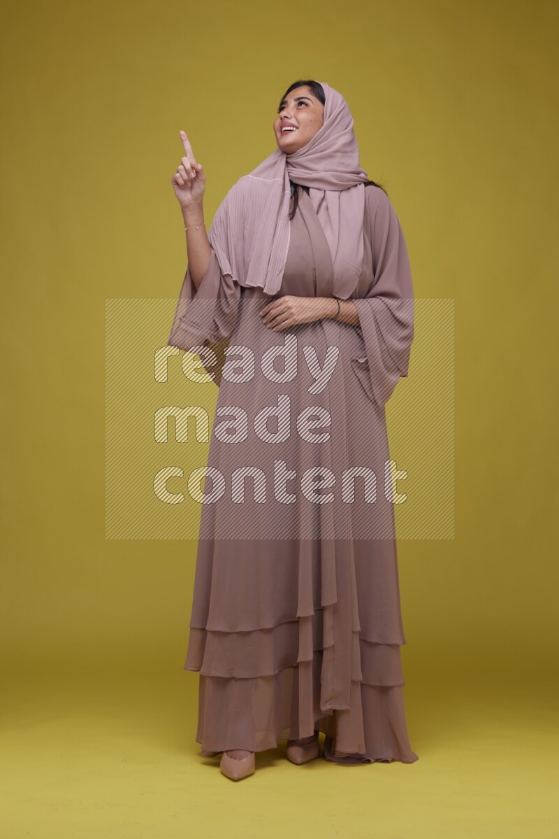 A woman Pointing on a Yellow Background wearing Brown Abaya with Hijab