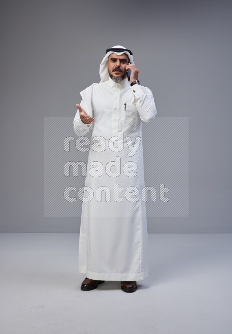 Saudi man Wearing Thob and white Shomag standing talking on phone on Gray background