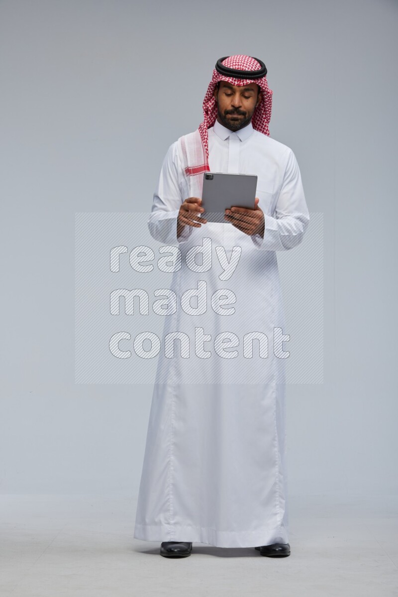 Saudi man Wearing Thob and shomag standing working on tablet on Gray background