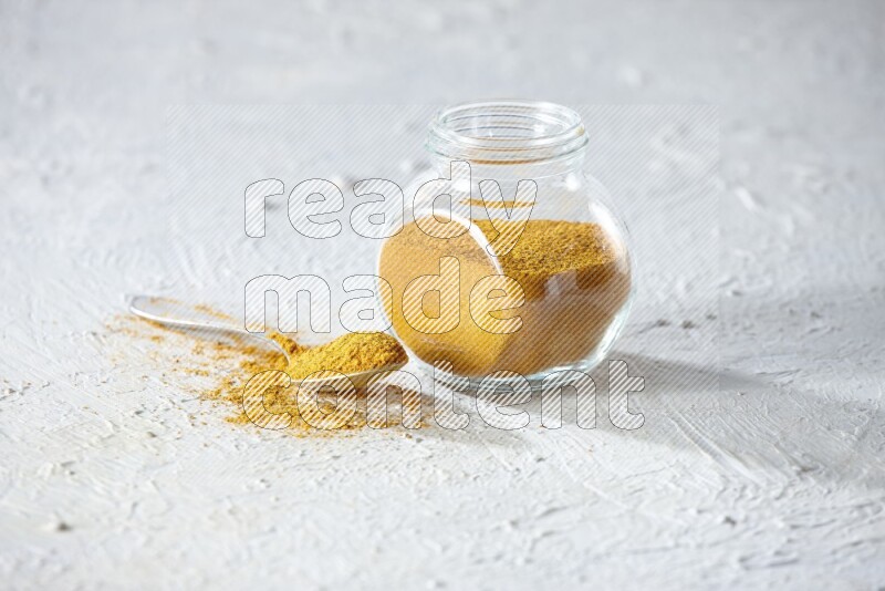 A glass spice jar and metal spoon full of turmeric powder on textured white flooring