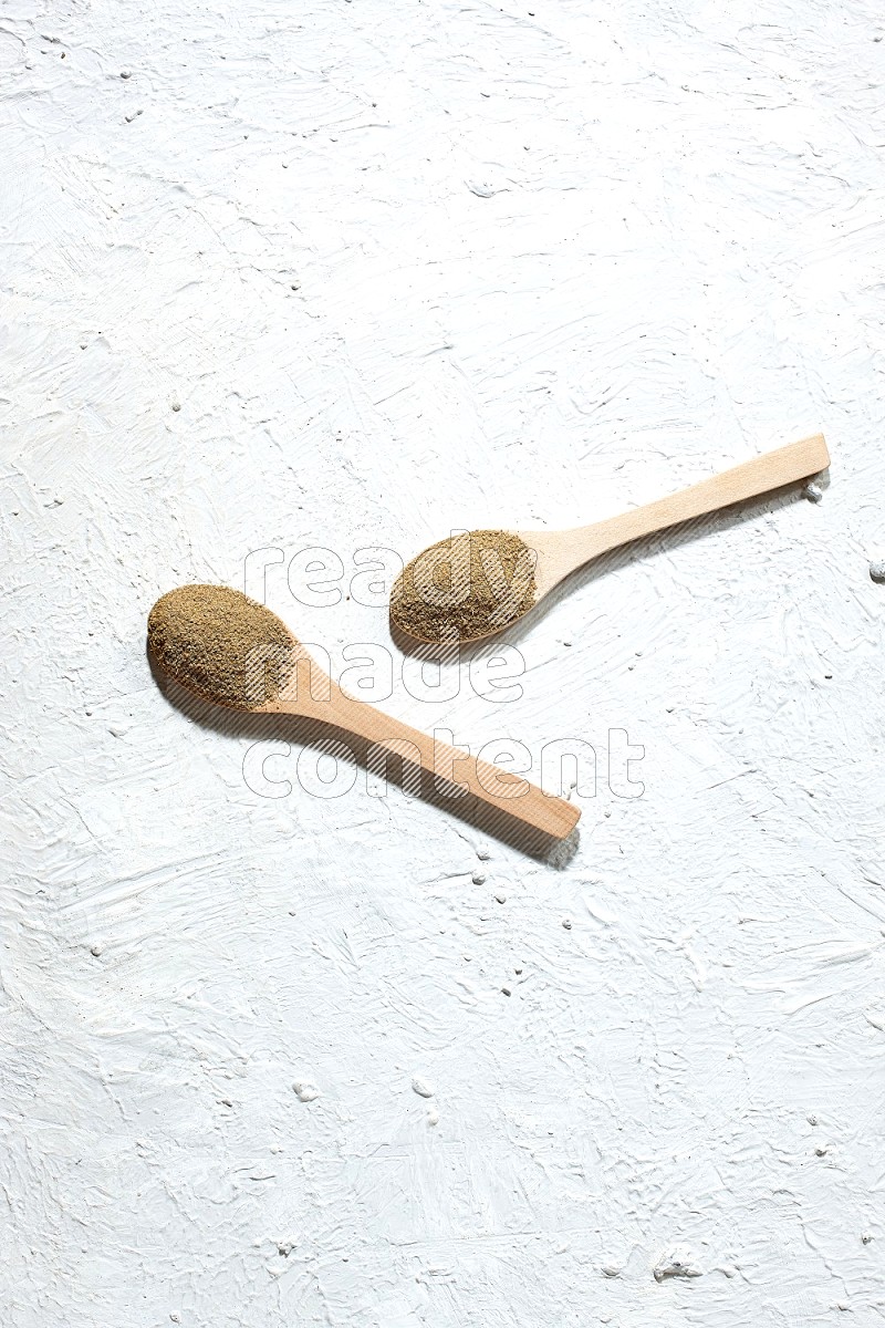 2 Wooden spoons full of cumin powder on a textured white flooring