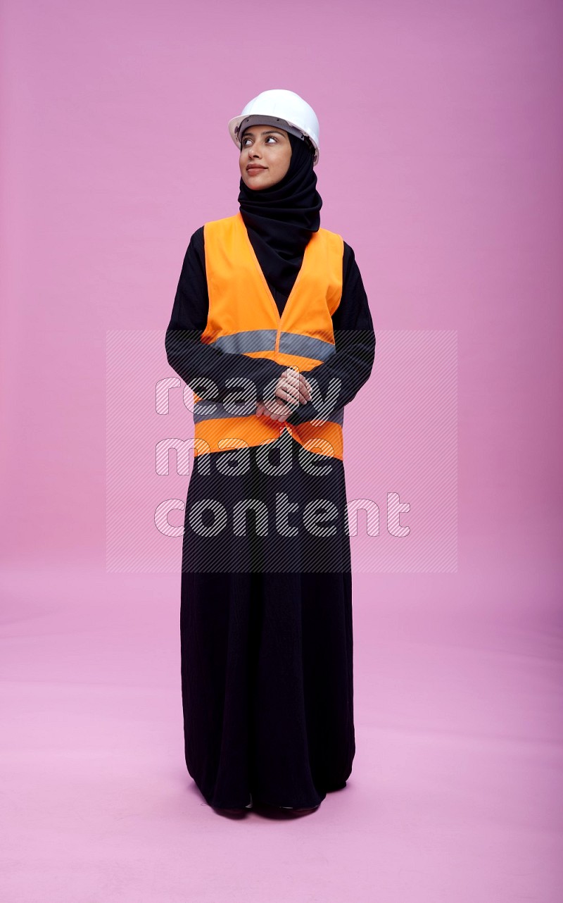 Saudi woman wearing Abaya with engineer vest and helmet standing interacting with the camera on pink background