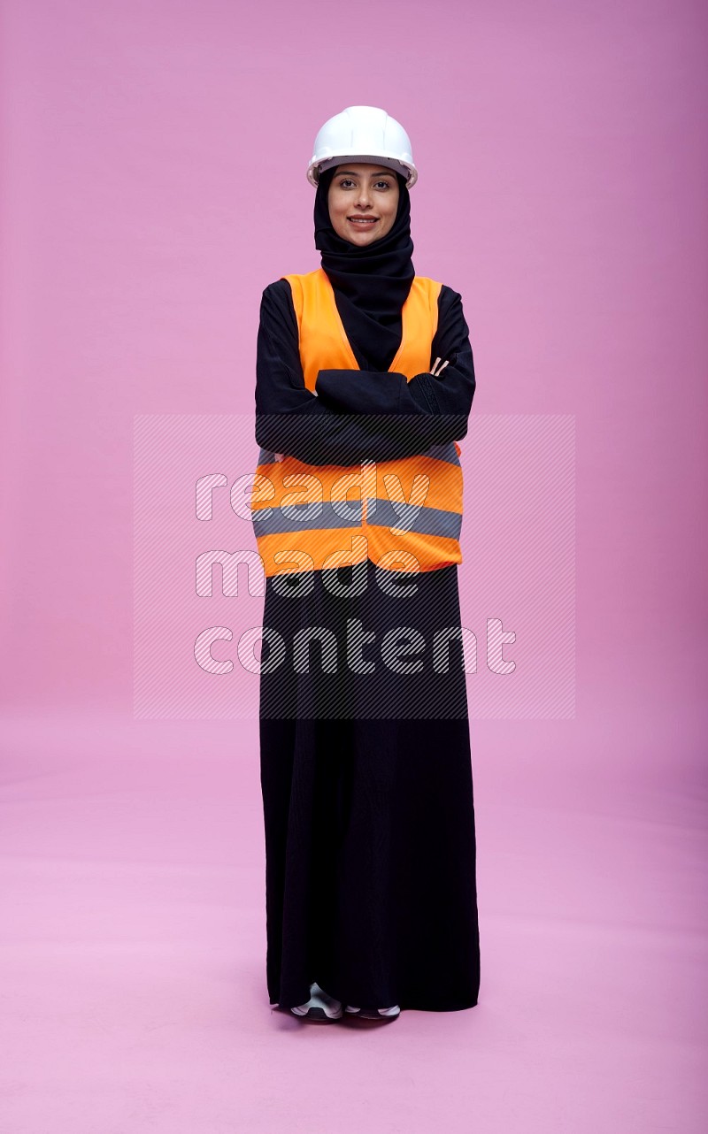 Saudi woman wearing Abaya with engineer vest and helmet standing with crossed arms on pink background