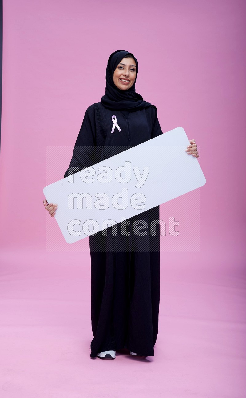 Saudi woman wearing pink ribbon on Abaya standing holding board on pink background