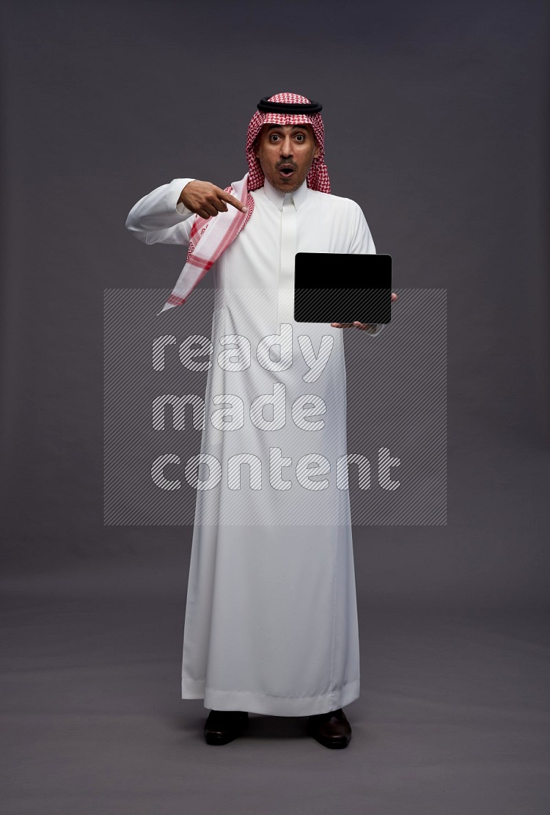Saudi man wearing thob and shomag standing showing tablet to camera on gray background