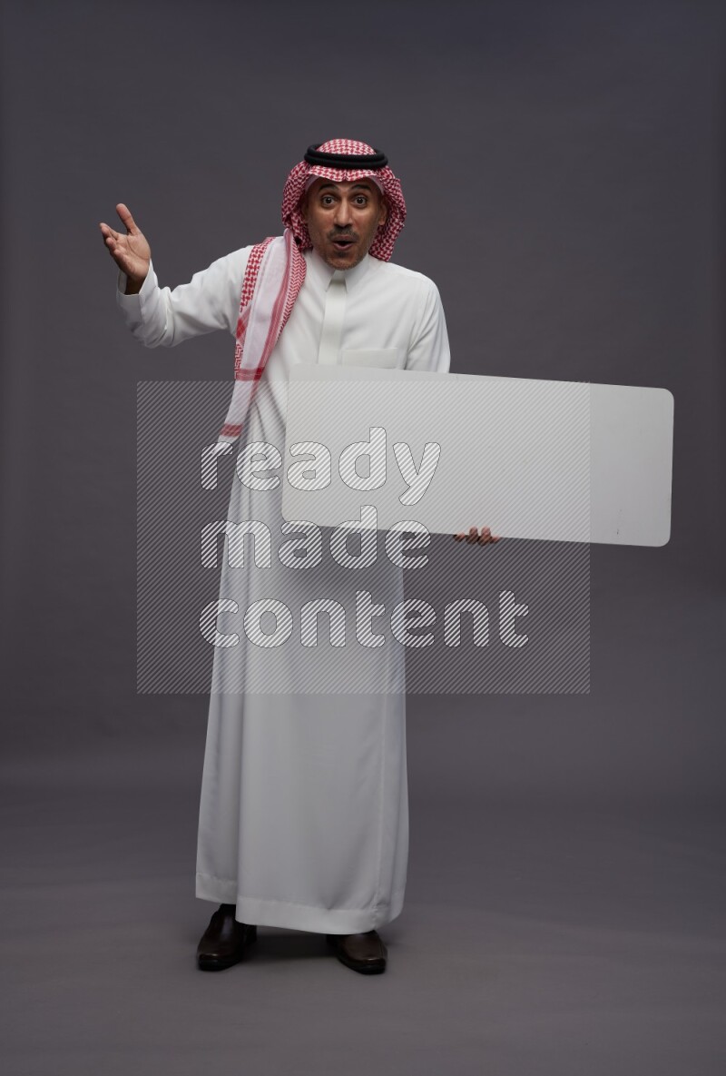 Saudi man wearing thob and shomag standing holding board on gray background