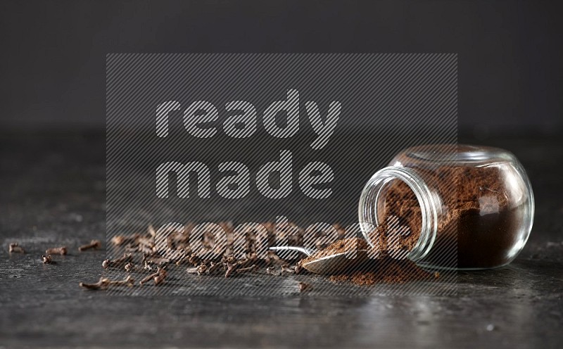 A flipped glass spice jar and a metal spoon full of cloves powder with cloves spread on textured black flooring