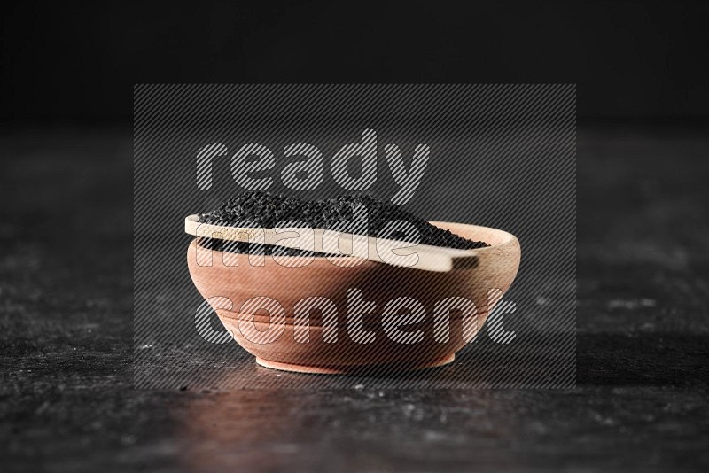 A wooden bowl full of black seeds with wooden spoon full of the seeds on it on a textured black flooring