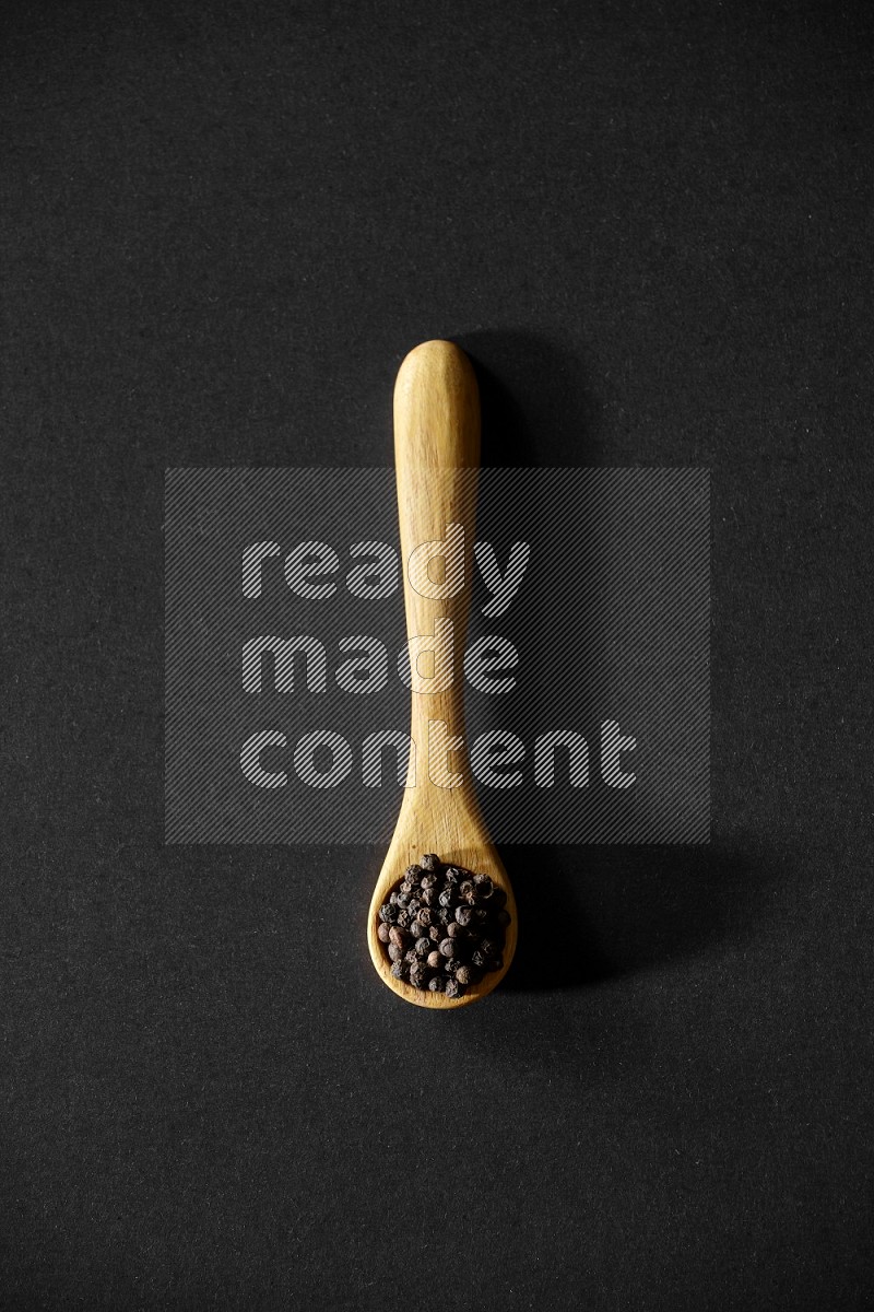 A wooden spoon full of black pepper on a black flooring
