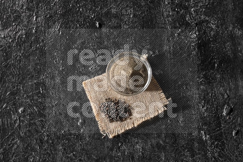 A glass bowl full of black pepper powder and black pepper beads on burlap fabric on textured black flooring