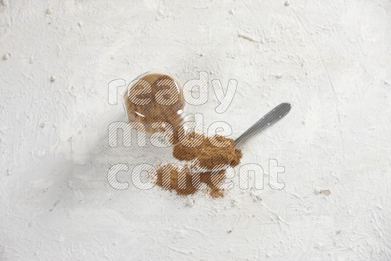 Flipped herbs glass jar full of cinnamon powder with a metal spoon full of powder on a textured white background