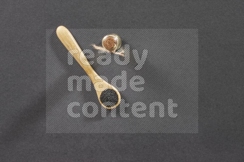 A wooden spoon full of black seeds and a glass bottle of black seeds oil on a black flooring
