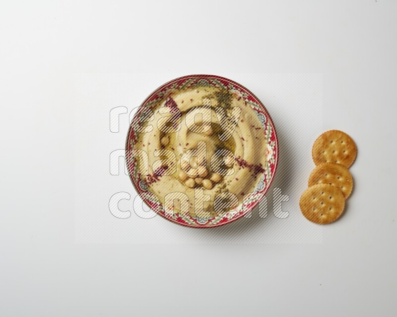 Hummus in a red plate with patterns garnished with zaatar & sumak on a white background