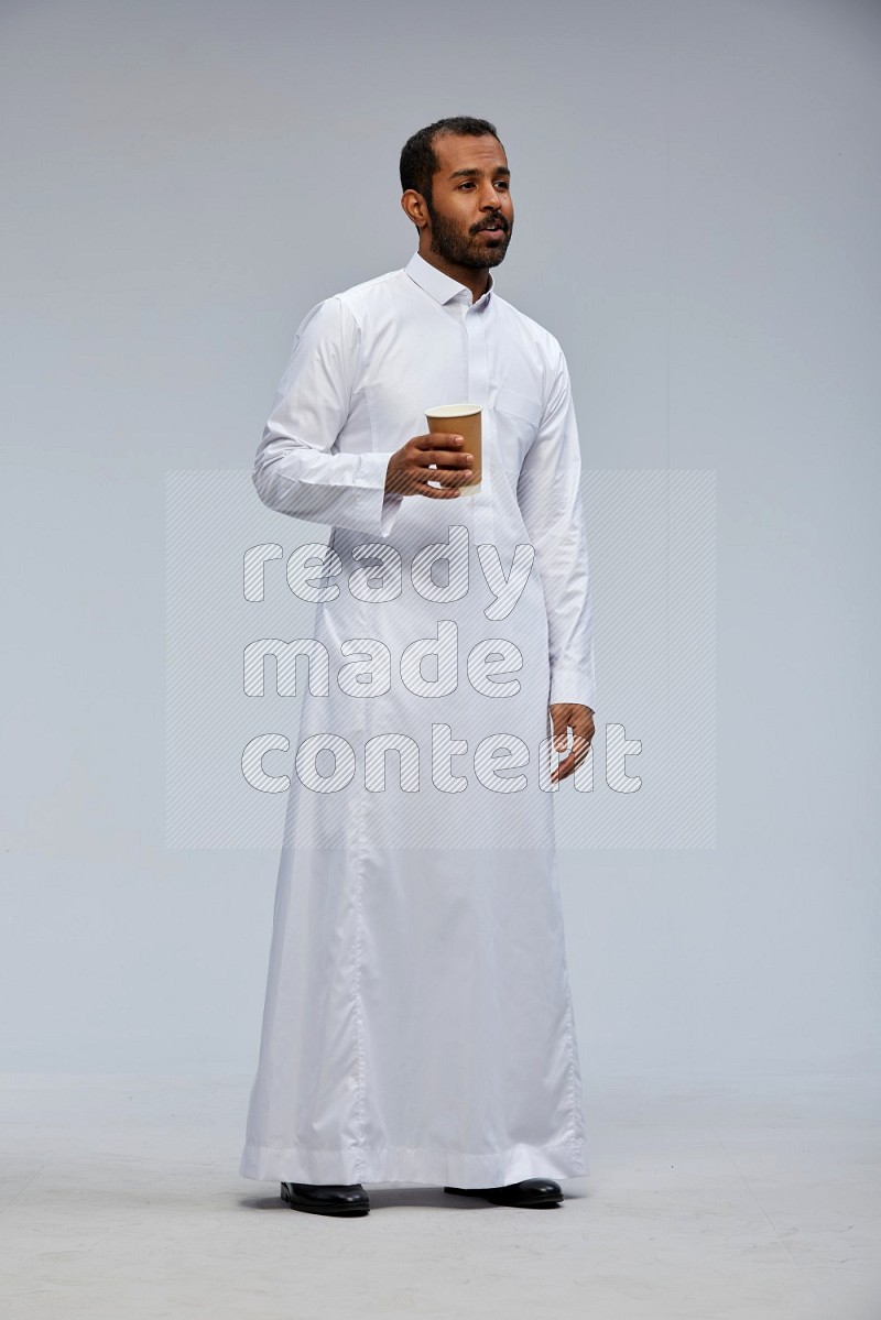 Saudi man wearing thob standing holding paper cup on gray background