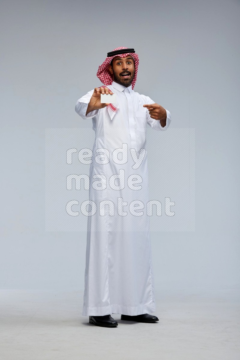 Saudi man wearing thob and shomag standing holding ATM card on gray background