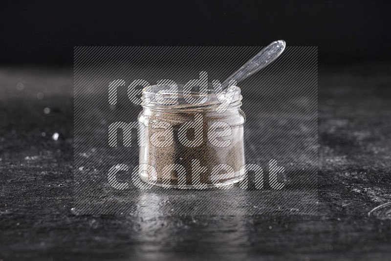 A glass jar full of black pepper powder and a metal spoon on a textured black flooring