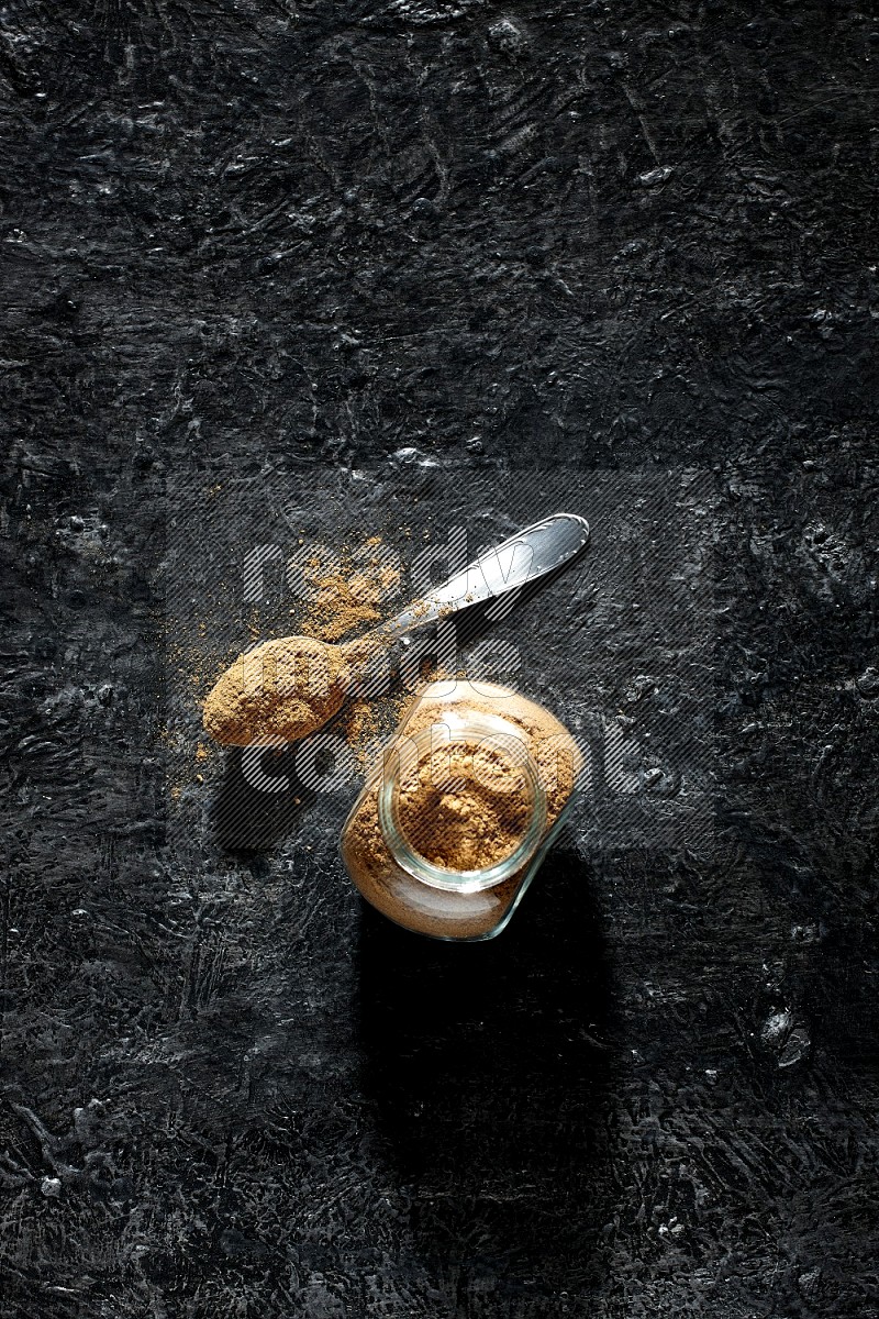 A glass spice jar and metal spoon full of allspice powder on a textured black flooring