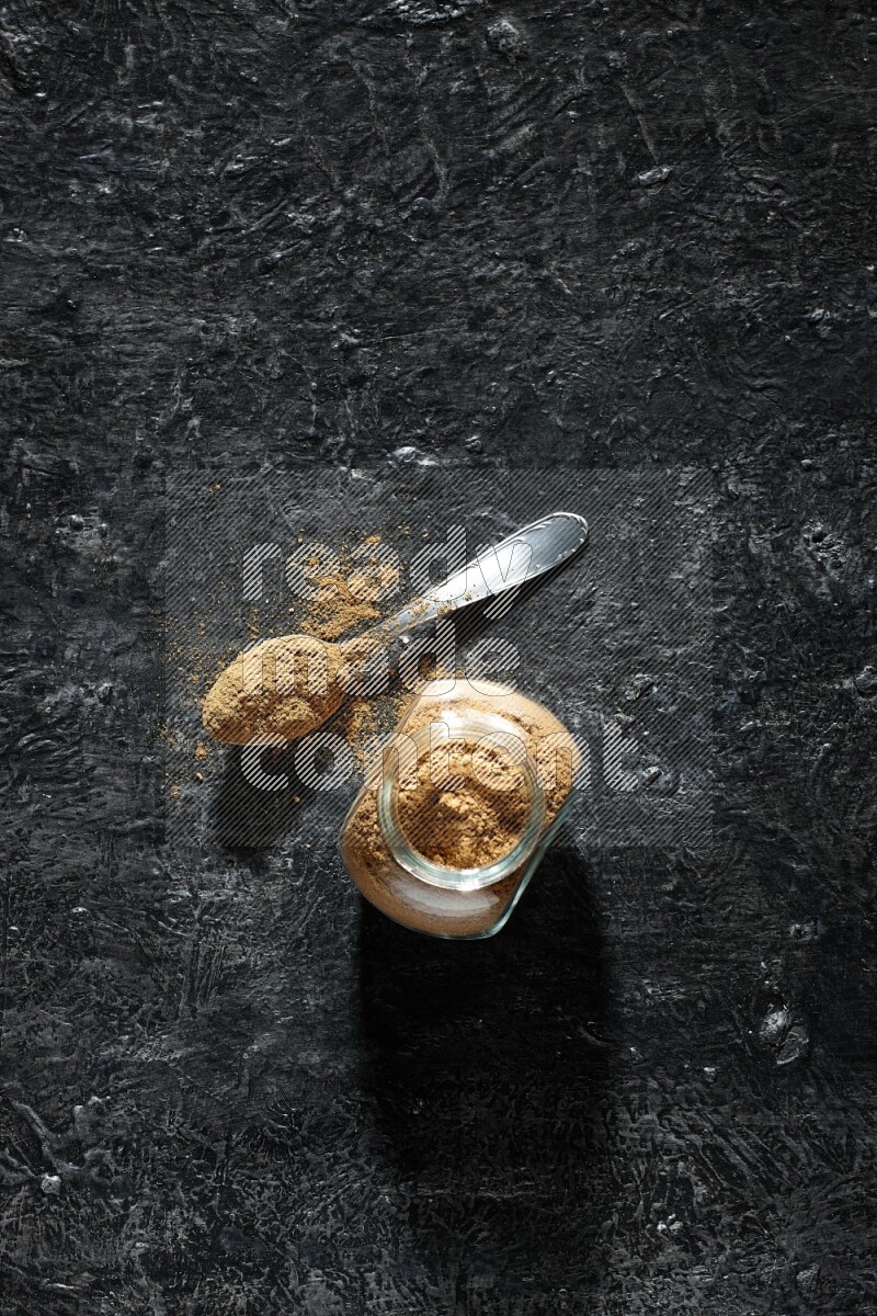 A glass spice jar and metal spoon full of allspice powder on a textured black flooring
