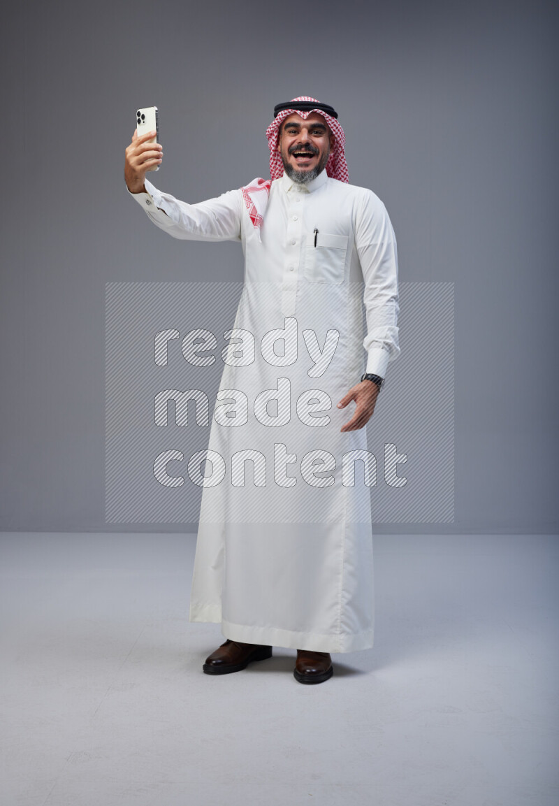 Saudi man Wearing Thob and red Shomag standing taking selfie on Gray background