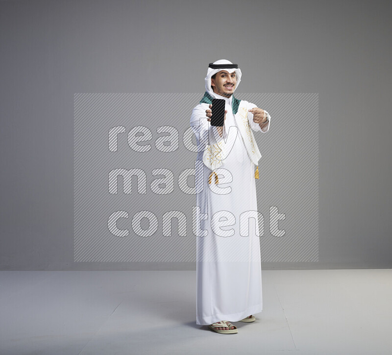 A Saudi man standing wearing thob and white shomag with flag scarf on his neak showing phone on gray background