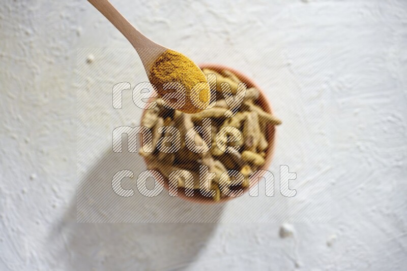 A wooden spoon full of turmeric powder above a wooden bowl full of dried turmeric whole fingers on a textured white flooring
