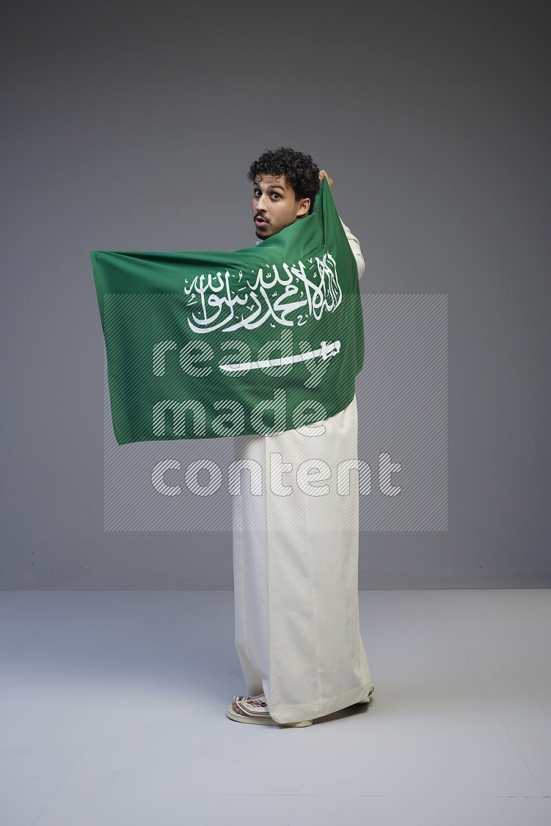 A Saudi man standing wearing thob holding big Saudi flag on gray background