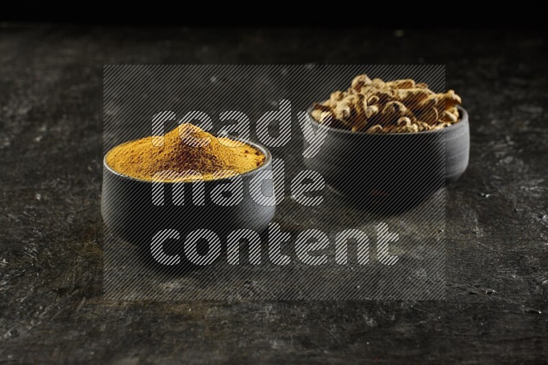 2 pottery black bowls, one full of turmeric powder and the other full of dried turmeric whole fingers on a textured black flooring