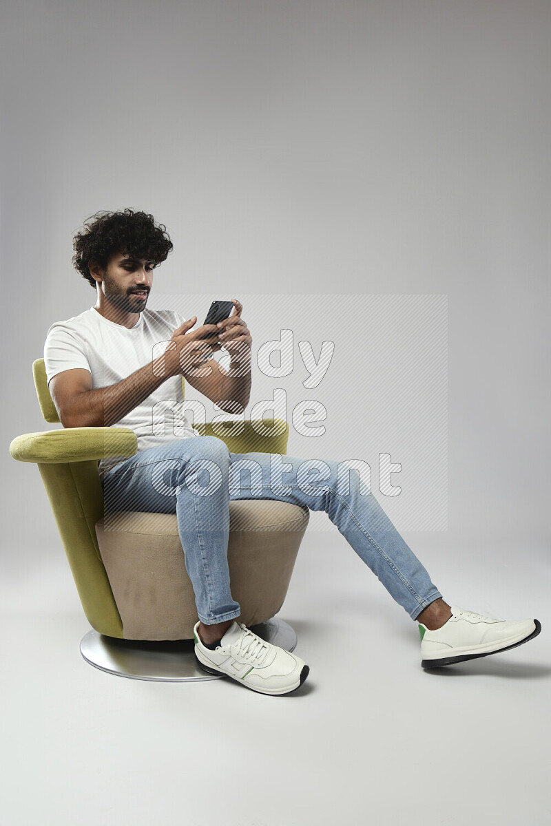 رجل يرتدي ملابس كاجوال يجلس علي كرسي و يرسل رسائل نصية علي الهاتف علي خلفية بيضاء