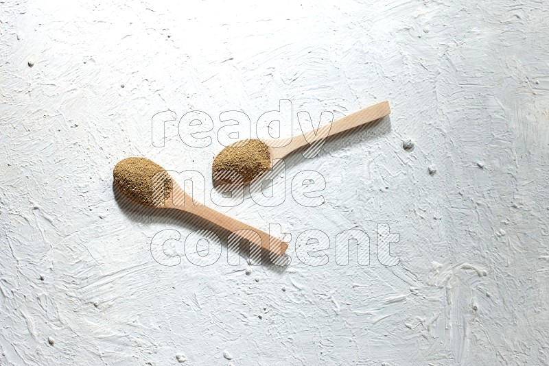 2 Wooden spoons full of cumin powder on a textured white flooring
