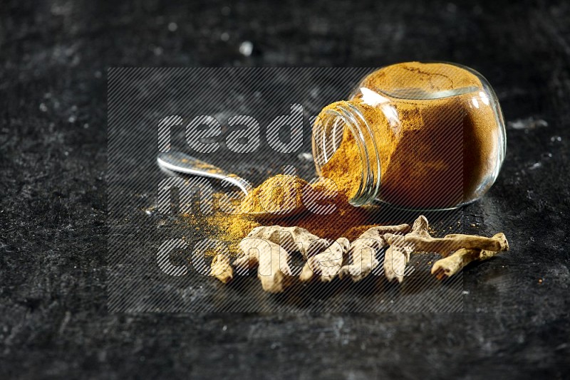 A flipped glass spice jar and metal spoon full of turmeric powder and powder spilled out of it with dried whole fingers on textured black flooring