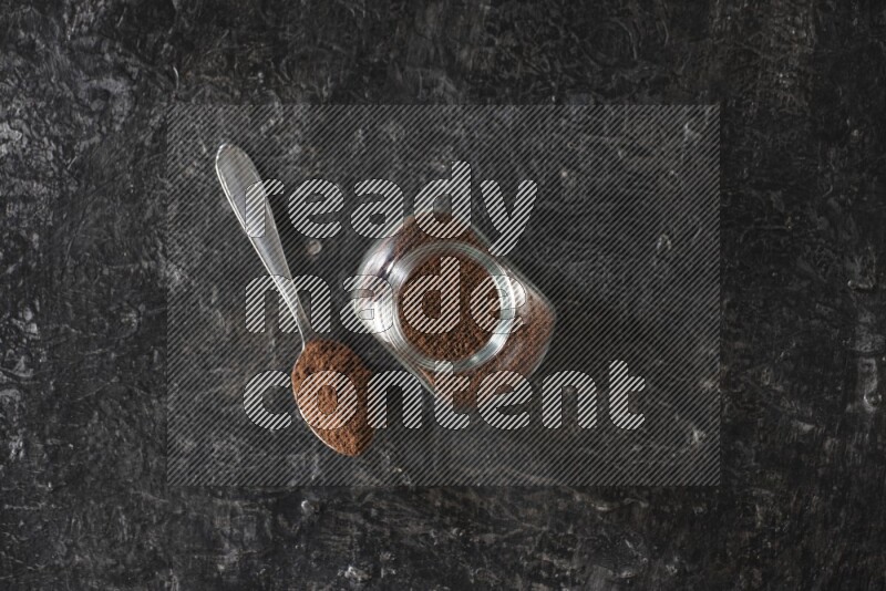 A glass spice jar and a metal spoon full of cloves powder on textured black flooring