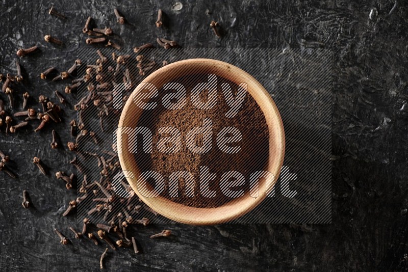 A wooden bowl full of cloves powder on a textured black flooring