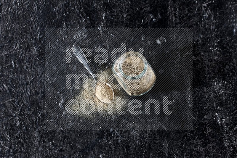 وعاء زجاجي وملعقة معدنية مملوءة بمسحوق الفلفل الأبيض على أرضية سوداء