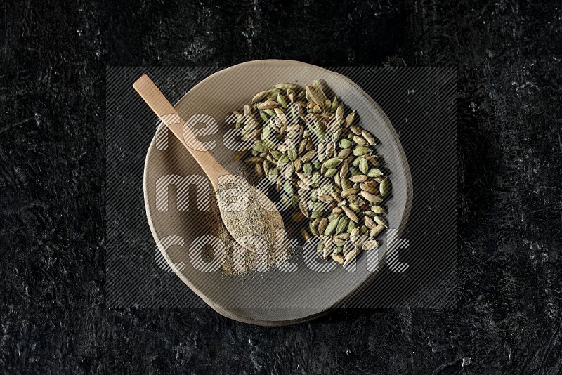 A plate filled with cardamom seeds and a wooden spoon full of cardamom powder on a textured black flooring