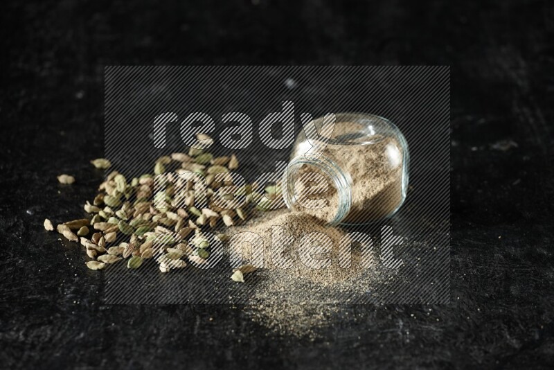 A flipped glass spice jar full of cardamom powder and powder spilled out of it with cardamom seeds spreaded on textured black flooring