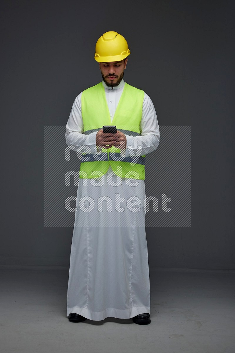 A Saudi man wearing Thobe with a yellow safety vest and white helmet standing and using his phone eye level on a gray background