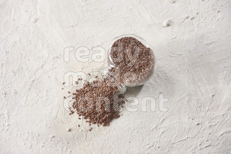 A glass spice jar full of flax seeds flipped and seeds spread out on a textured white flooring