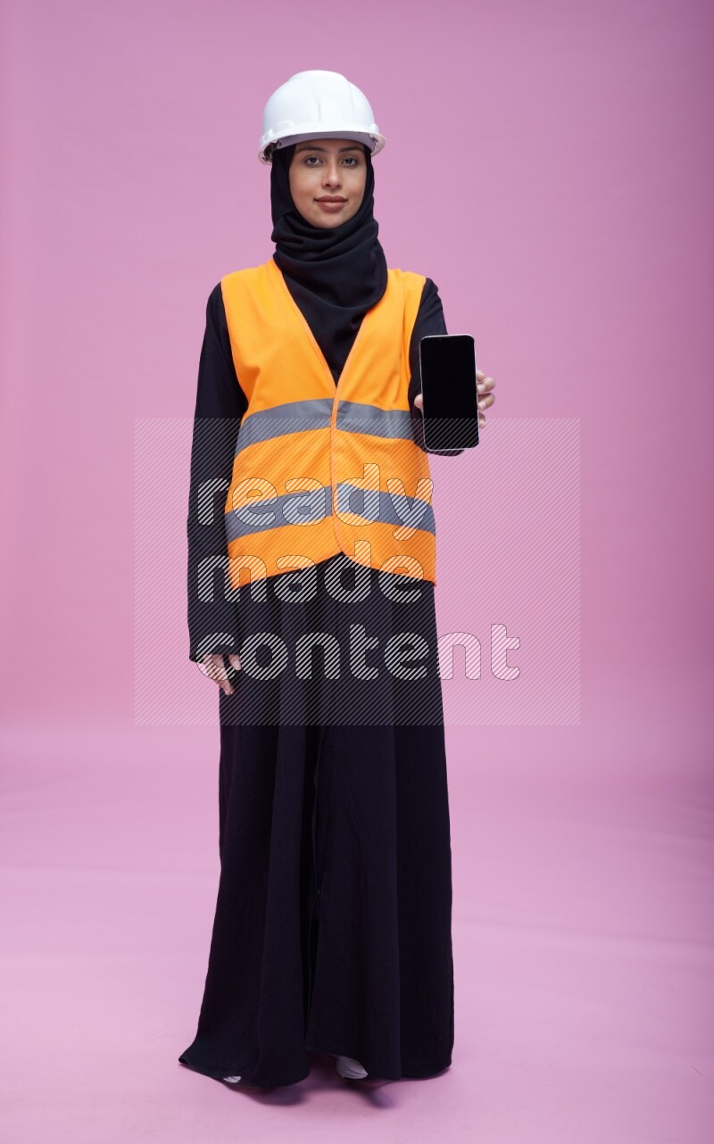 Saudi woman wearing Abaya with engineer vest and helmet standing showing phone to camera on pink background