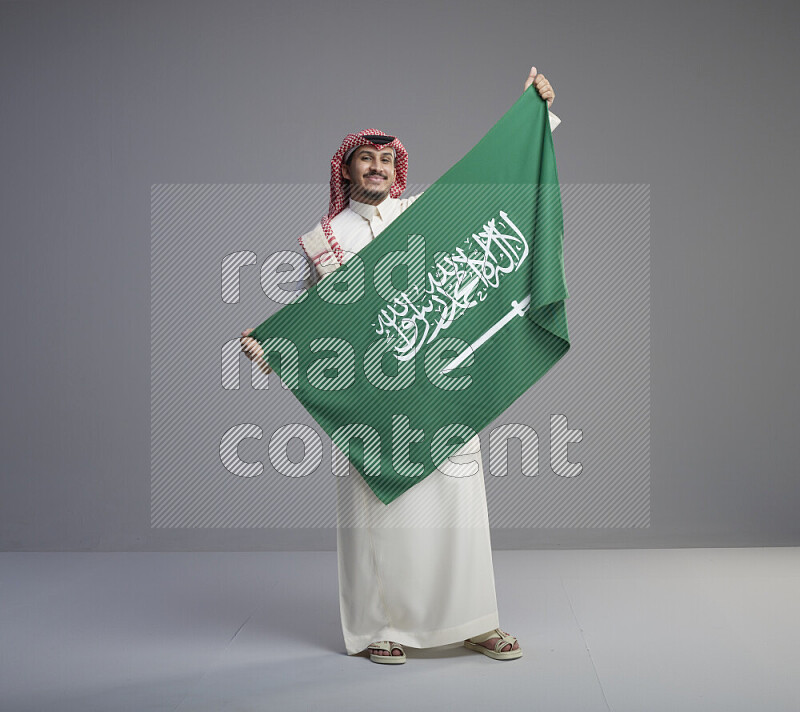 A saudi man standing wearing thob and red shomag holding big saudi flag on gray background