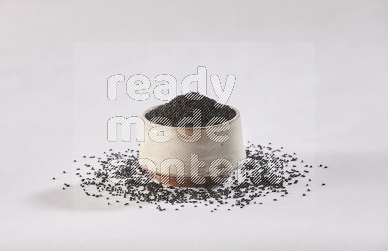A beige pottery bowl full of black seeds and more seeds spread on a white flooring