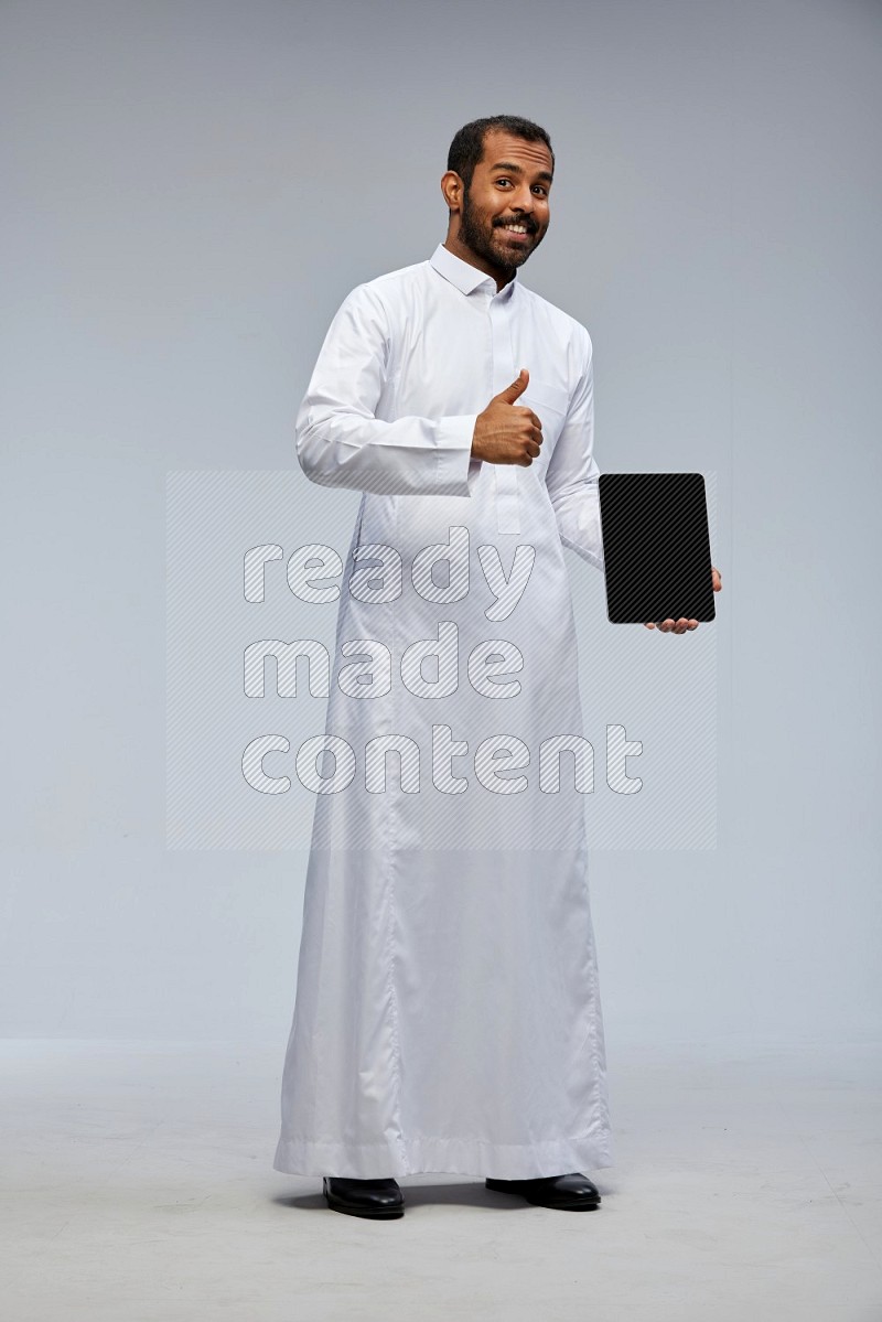 Saudi man wearing Thob standing showing tablet to camera on Gray background