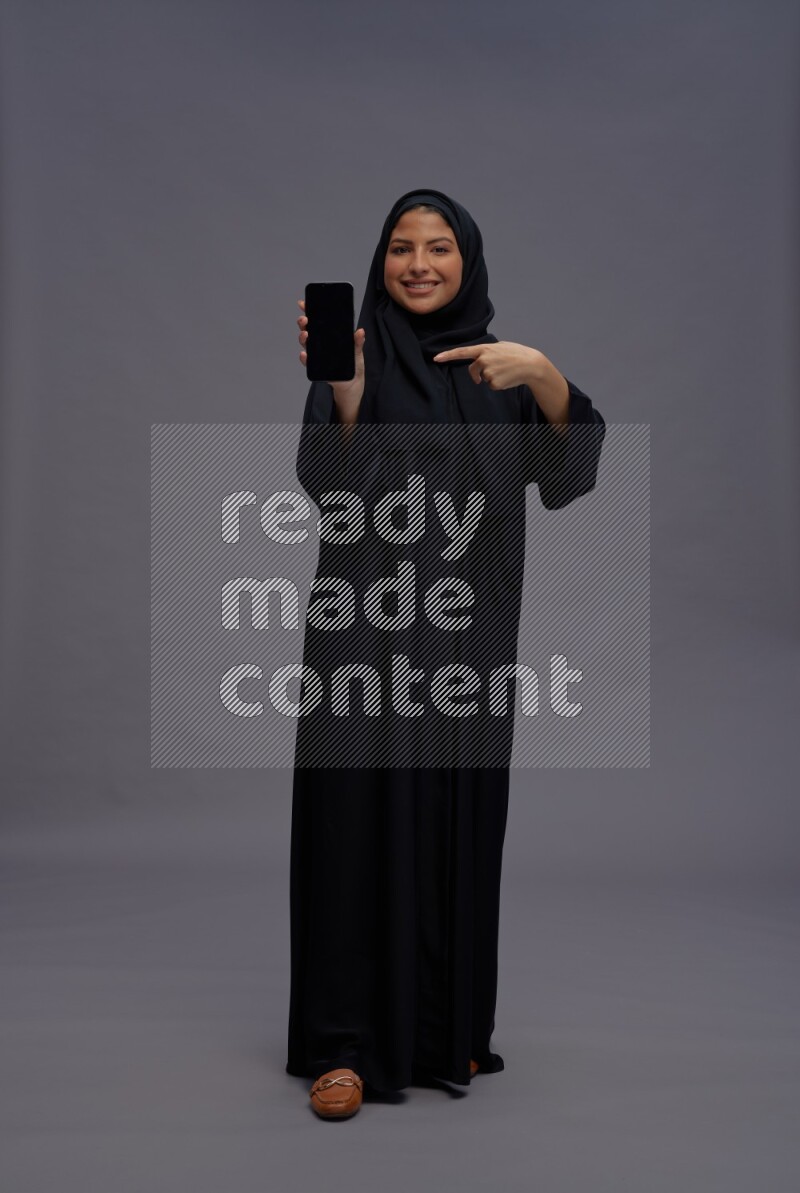 Saudi woman wearing Abaya standing showing phone to camera on gray background